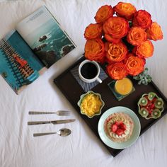 an arrangement of food and drink on a tray with a book, spoons, fork and knife