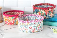 three flowered fabric baskets sitting on top of a counter next to scissors and thread