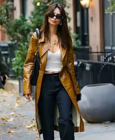 a woman walking down the street wearing black pants and a brown leather trench coat with white top