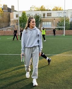 a woman walking across a soccer field with other people on the sidelines behind her