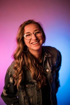 a woman with glasses smiling at the camera in front of a blue and pink background