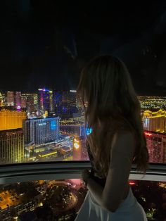 a woman standing in front of a window looking out at the city lights and buildings