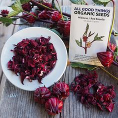 a bowl of red flowers next to an organic seed packet on a wooden table with leaves