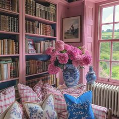 pink flowers are in a blue and white vase next to pillows on a couch with bookshelves