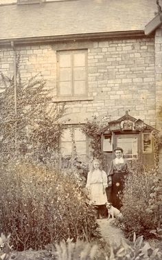 an old photo of two people standing in front of a house with bushes and flowers