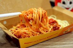 someone is eating spaghetti with chopsticks in a cardboard box on a wooden table