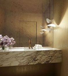 a bathroom with marble counter top and lights on the wall, along with purple flowers