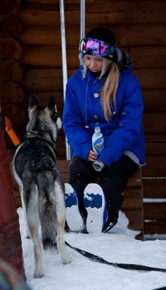 a woman sitting next to a dog in the snow with a bottle of water on her lap