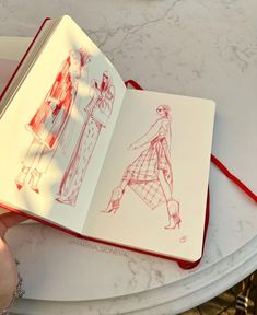 a hand holding an open book with drawings on it and a red string attached to the cover