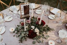 the table is set with white plates and silverware, red flowers, greenery and candles