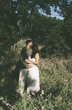 a man and woman standing in the middle of a field with wildflowers behind them