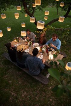 a group of people sitting at a table with paper lanterns hanging from the trees in the background