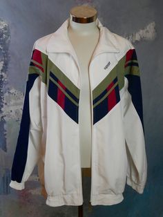 "This 1990s European windbreaker jacket is white, but has a wide band of blue, red, and taupe green across the chest and down the sleeves. The jacket zips closed in front, and you can leave it unzipped a little bit to create a wing collar or zip it all the way up to create a high band collar. The jacket has two zippered pockets on the front, and is lined in a cooling polyester mesh fabric. Brand label: Model Paris Collection Size: XXXL (54 to 56 US/UK) Material: Polyester Condition: Excellent (c 90s Style White Outerwear For Outdoor, 90s Style White Track Jacket For Outdoor, White Retro Windbreaker For Outdoor, White 90s Style Track Jacket For Outdoor, Vintage White Windbreaker For Fall, 90s Style White Long Sleeve Track Jacket, Retro White Track Jacket For Outdoor, White 90s Track Jacket With Long Sleeves, White 90s Track Jacket For Outdoor