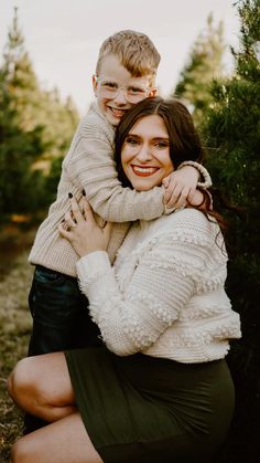 a woman holding a boy in her arms and smiling at the camera with trees in the background