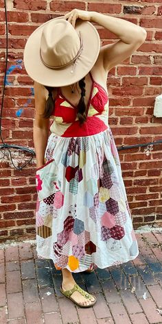 a woman in a dress and hat standing by a brick wall with her hands on her head