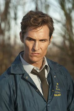 a man in a blue jacket and tie looking off to the side with trees in the background