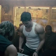 a man in white tank top standing next to a table with food on it and people around him