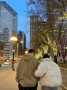 two people are walking down the sidewalk in front of tall buildings and trees with lights on them