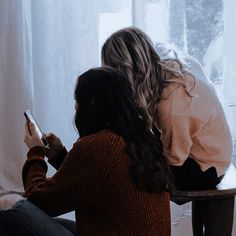 two women sitting on the floor looking at their cell phones in front of a window