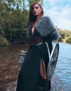 a woman in a long black dress and fur coat standing next to a body of water