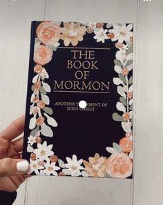 the book of mormon is being held up in front of a white wooden wall with flowers on it