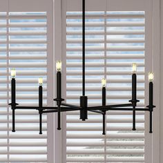 a black chandelier with five lit candles hanging from it's center, in front of white shutters