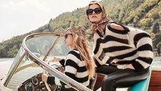 two women in black and white sweaters sitting on the back of a boat with mountains in the background