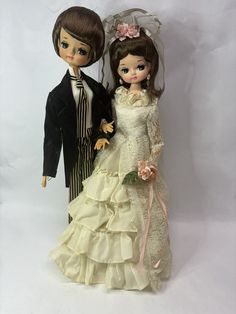 two dolls dressed in wedding attire standing next to each other on a white background with one holding the bride's hand