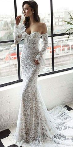 a woman standing in front of a window wearing a white wedding dress with long sleeves