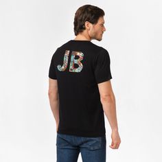 Man wearing a black t-shirt with large, multicolored 'JB' letters on the back. The letters are composed of various patterns and designs. The man is shown from the back, standing with his head turned to the side, wearing blue jeans. Black Crew Neck T-shirt With Front Print, Black Crew Neck Top With Back Print, Black Crew Neck Top With Front Print, Casual Crew Neck T-shirt With Back Print, Casual Crew Neck Top With Back Print, Casual Black T-shirt With Front Print, Casual Black Relaxed Fit T-shirt, Casual Black T-shirt With Relaxed Fit, Black Casual Pre-shrunk T-shirt