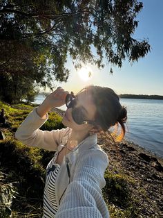 a woman standing on top of a grass covered field next to the ocean drinking from a bottle