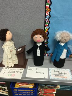 three dolls are sitting on a table in front of a bulletin board and bookshelf