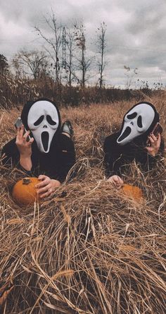 two people with masks on their faces sitting in the grass holding pumpkins and talking on cell phones