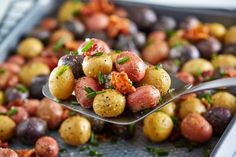 a spoon full of cooked potatoes and olives on a baking sheet with other vegetables in the background