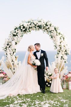 a bride and groom standing in front of an archway with white flowers on the grass