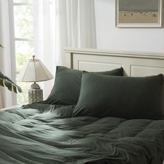 a bed with green sheets and pillows next to a lamp on a table in front of a window