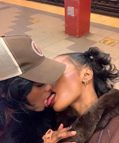 two women are kissing each other on the subway platform, one is wearing a hat and the other has her mouth open