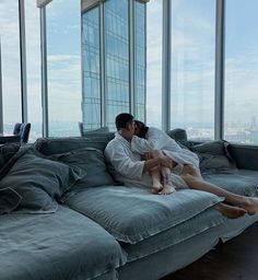 a man and woman sitting on top of a couch in front of large glass windows