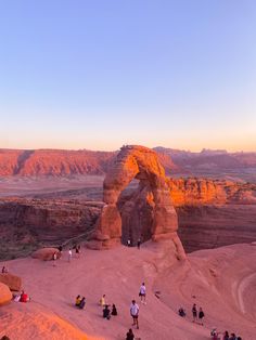 people are standing in the sand at sunset near an arch shaped rock formation, with mountains in the background