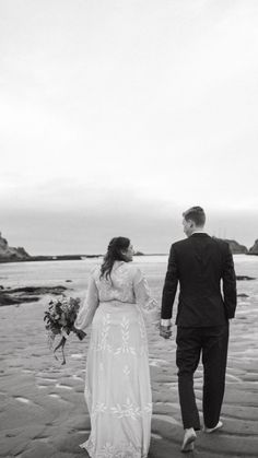 a bride and groom walking on the beach holding hands with flowers in their hand, black and white photo