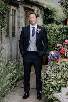 a man wearing a suit and tie standing in front of some flowers with his hands in his pockets