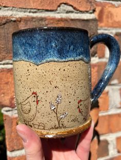 a hand holding a blue and brown coffee cup with birds painted on the side, in front of a brick wall