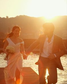 a bride and groom are walking on the dock holding hands as the sun sets behind them