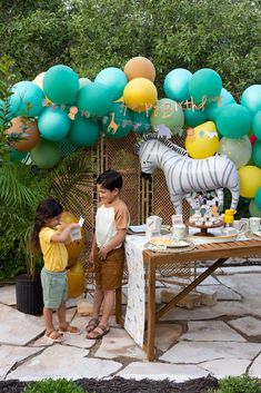 two children standing in front of a table with cake and balloons on it at a zoo themed birthday party