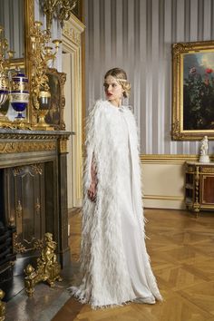 a woman standing in front of a fire place wearing a white feathered gown and cape