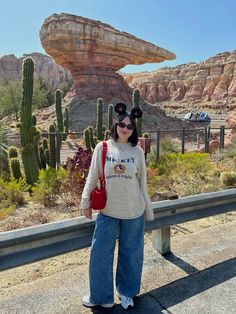 a woman standing on the side of a road with cactus in the background and a rock formation behind her