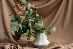 a white vase filled with lots of greenery sitting on top of a bed next to a brown sheet