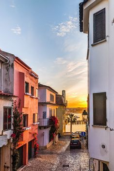 the sun is setting over some buildings and parked cars on the cobblestone street