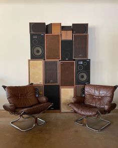 two brown leather chairs sitting next to each other in front of a wall with speakers on it