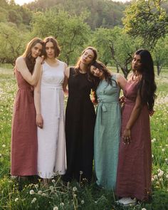 four women in long dresses posing for a photo on the grass with trees in the background